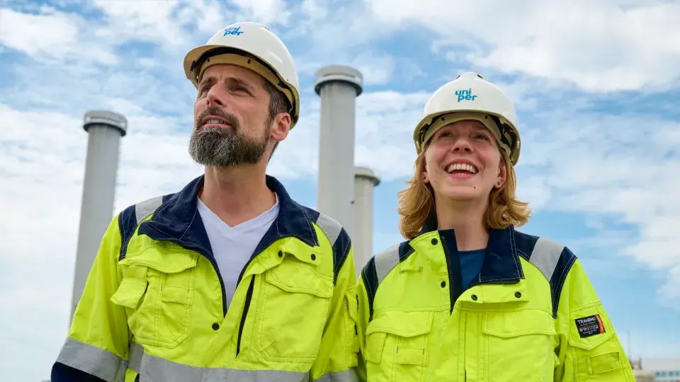 Two Uniper workers in high-visibility clothing and safety helmets standing outdoors, smiling, with industrial chimneys in the background.
