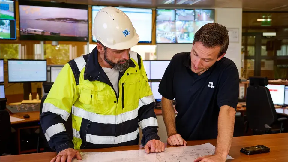Two Uniper employees discussing plans over a blueprint in a control room with multiple monitors.