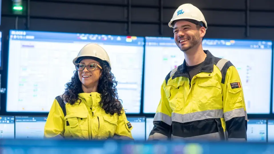 Two Uniper employees in safety gear smiling in a control room with screens in the background