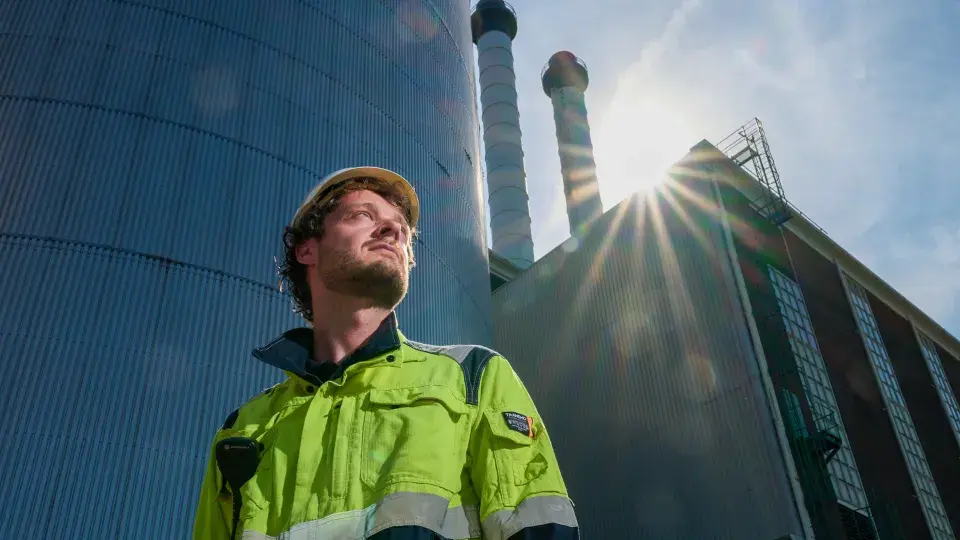 A worker in high-visibility gear and a hard hat stands confidently in front of an industrial facility with sunlight streaming past large chimneys.