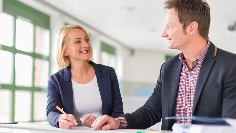 Two business professionals in a bright office discussing plans, with one holding a pen and smiling at the other.