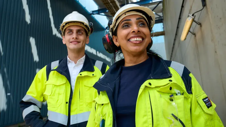 Two workers in high-visibility jackets and helmets, smiling while standing outdoors near an industrial facility.