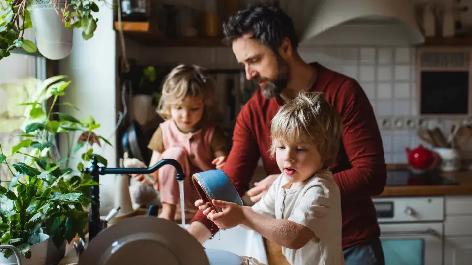 A father and his two young children washing dishes together in a cozy, plant-filled kitchen, symbolizing sustainable living and family care.