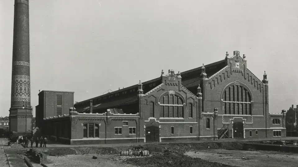 A historical black-and-white photo of a large brick factory building with a distinctive tall, patterned chimney and several workers standing in front of it.