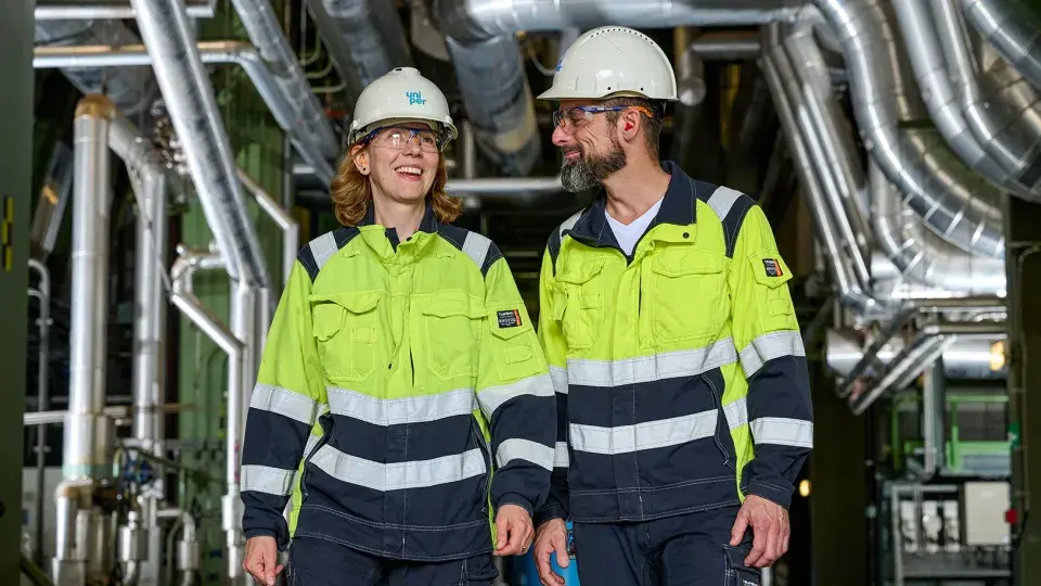 Two Uniper employees, a man and a woman, walking together inside a facility. They are smiling and wearing safety helmets and yellow work jackets with reflective strips.
