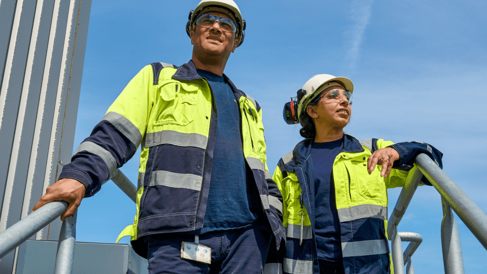 Two industrial workers in high-visibility safety jackets and helmets standing on a metal staircase, looking forward with a clear blue sky in the background