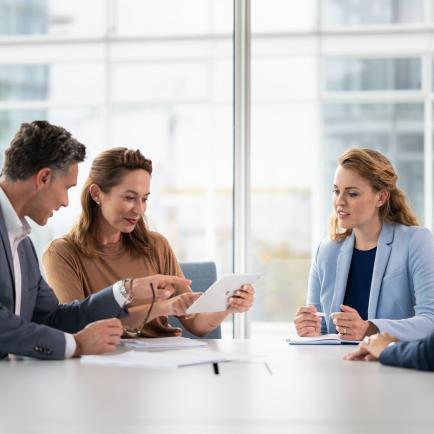 A man and three women in business outfits sit in a meeting room and talk to each other