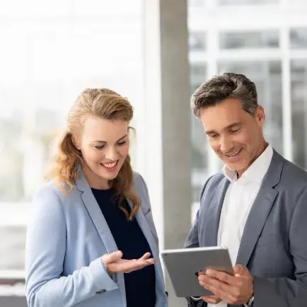 Two professionals in business attire discussing something on a tablet, smiling in a bright office environment.