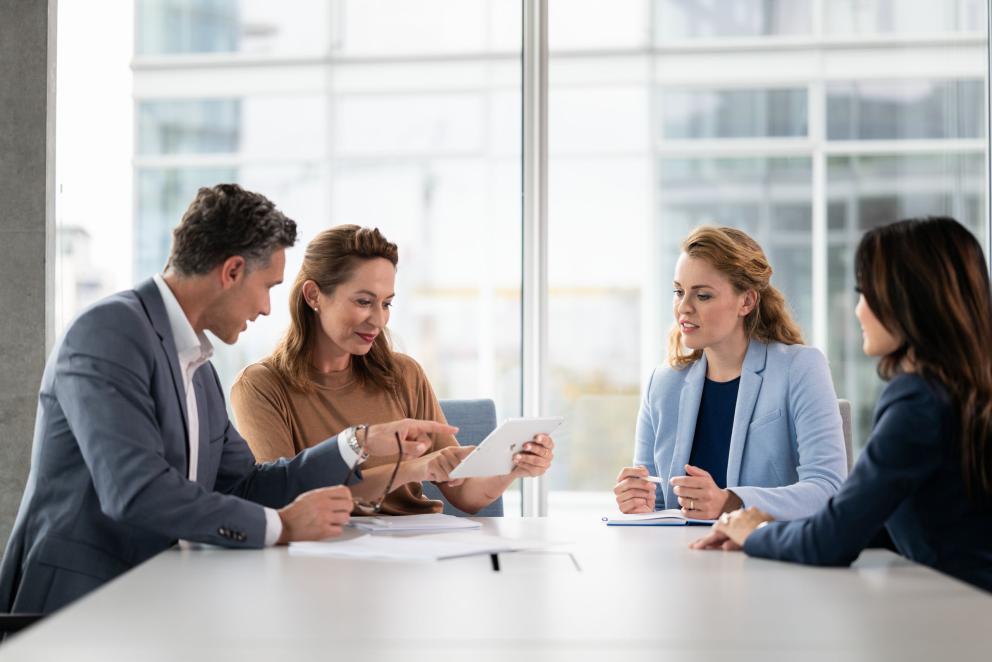 A man and three women in business outfits sit in a meeting room and talk to each other