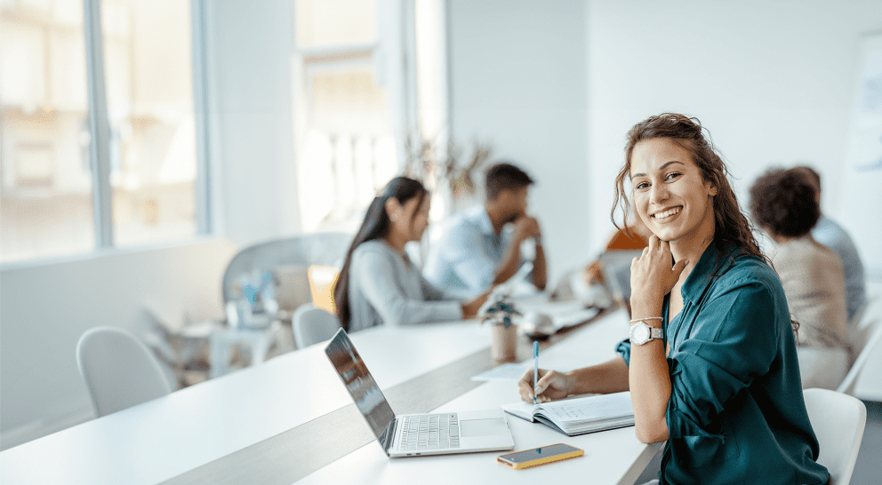 Female employee with laptop smiling at a meeting table, other employees in the background