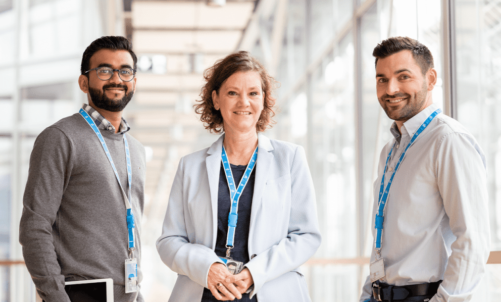 Three professionals wearing ID lanyards, standing and smiling in a bright, modern office hallway.