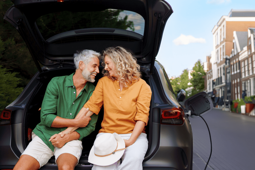 Smiling middle-aged couple sitting at the open trunk of an electric car, with a charging cable connected, sharing a warm moment in a cozy, urban setting.