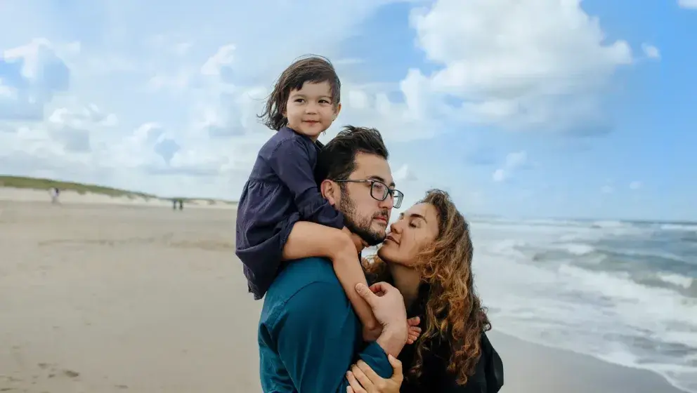 A family standing on a beach, with a young child on the father's shoulders and the mother kissing the father's cheek, as they enjoy a day by the sea.