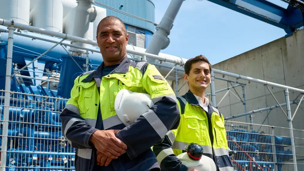 Two workers in high-visibility jackets standing outside at an industrial site, holding hard hats and smiling, with large equipment in the background