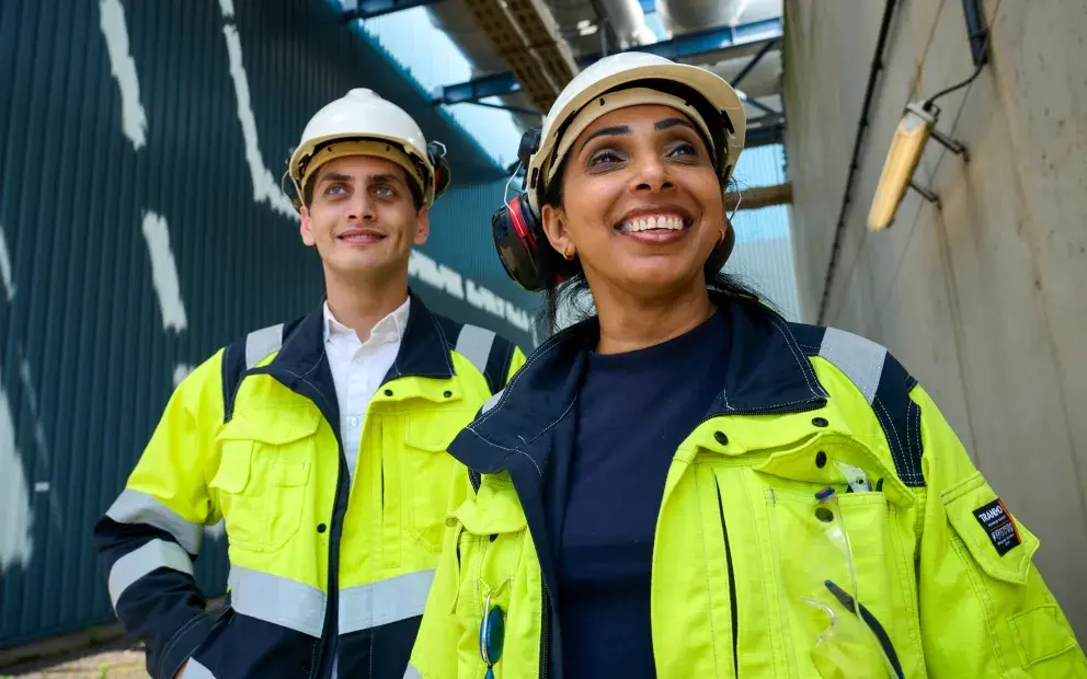 Two workers in high-visibility jackets and helmets, smiling while standing outdoors near an industrial facility.