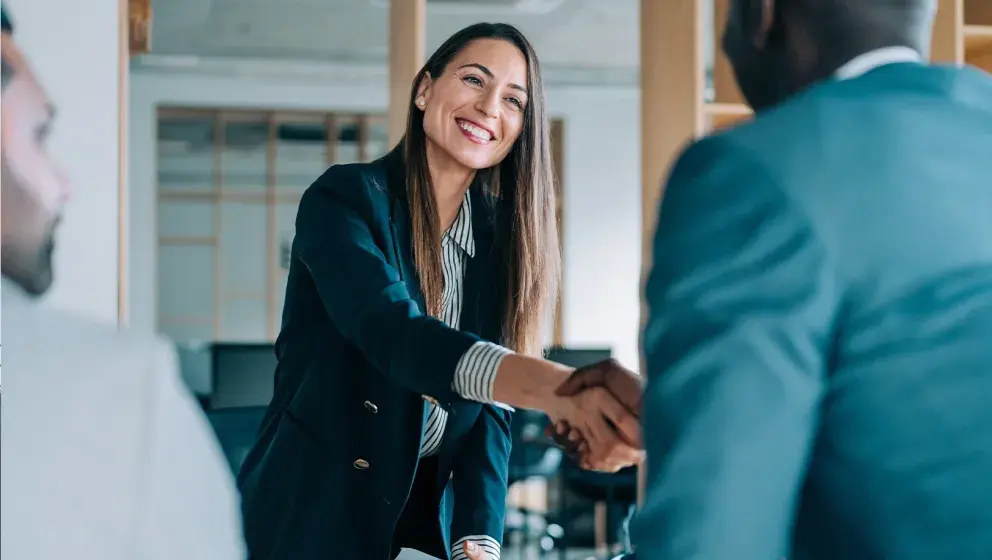 A woman in a suit smiling while shaking hands with a business partner in a modern office setting.