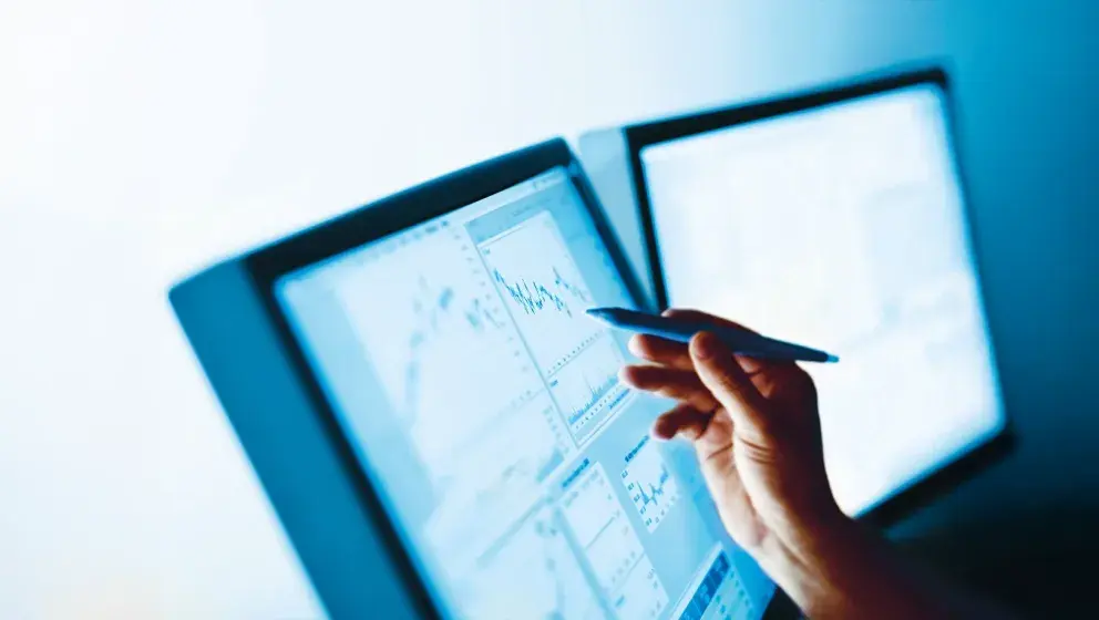 A hand holding a pen and pointing at financial data displayed on computer screens showing graphs and charts