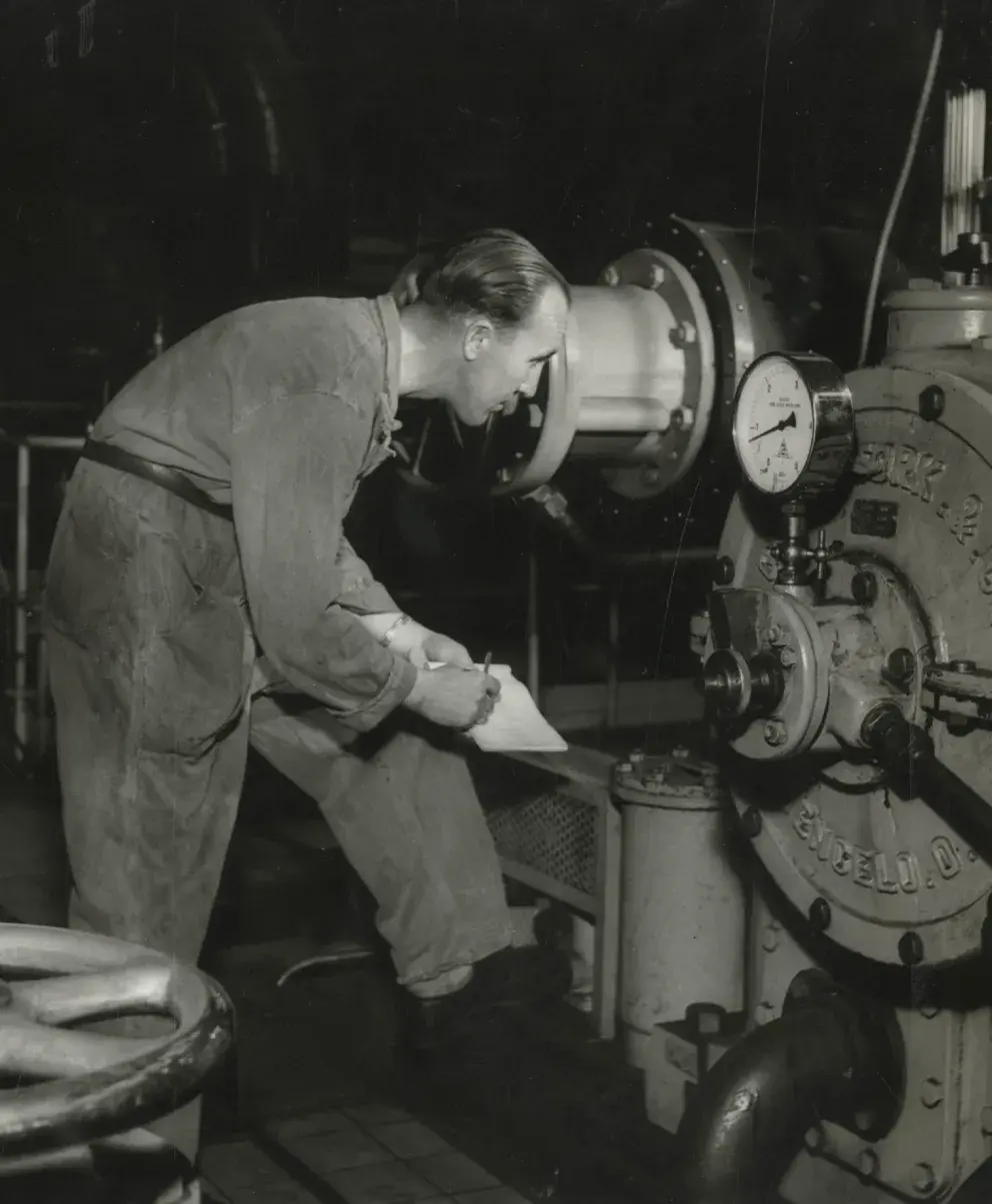 A historical black-and-white photo of a worker in a factory, checking a machine and taking notes.