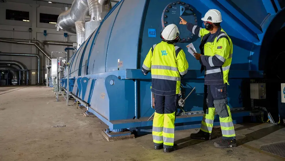 Two Uniper employees in high-visibility uniforms and helmets inspecting industrial machinery inside a power plant, pointing at a gauge on a large blue turbine.