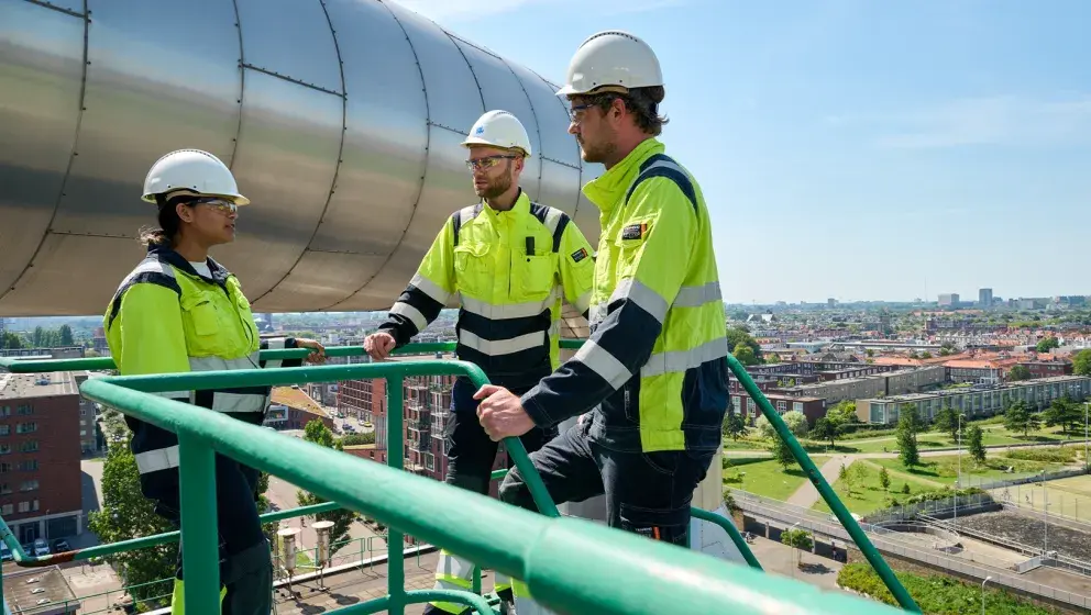 Three Uniper employees in high-visibility uniforms and helmets standing on a platform overlooking The Hague, with urban buildings and green spaces in the background.