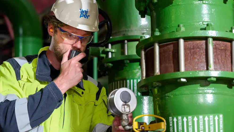 Uniper employee in a high-visibility uniform and helmet communicating via a radio device, inspecting green industrial pipes inside the power plant
