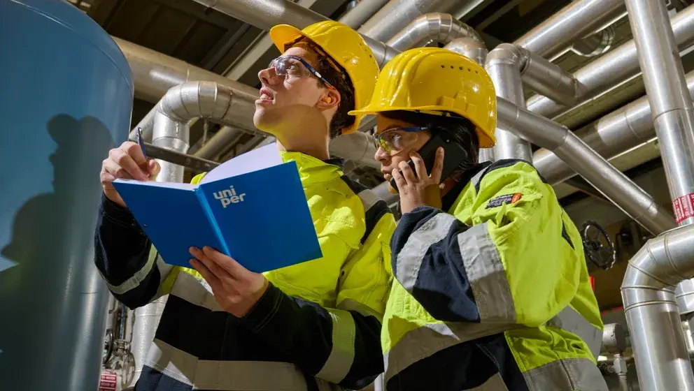 Two Uniper employees in high-visibility uniforms and yellow helmets working in an industrial setting, one holding a Uniper folder and pen, the other on a phone.