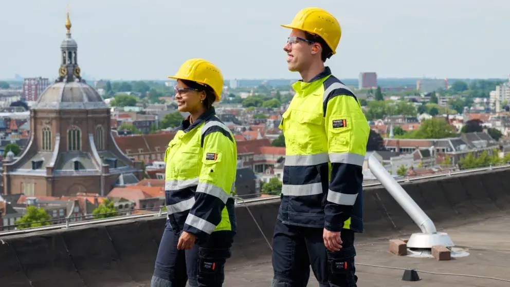 Two Uniper employees in high-visibility uniforms and yellow helmets walking on a rooftop with a view of Leiden's historic skyline, including a prominent church dome.