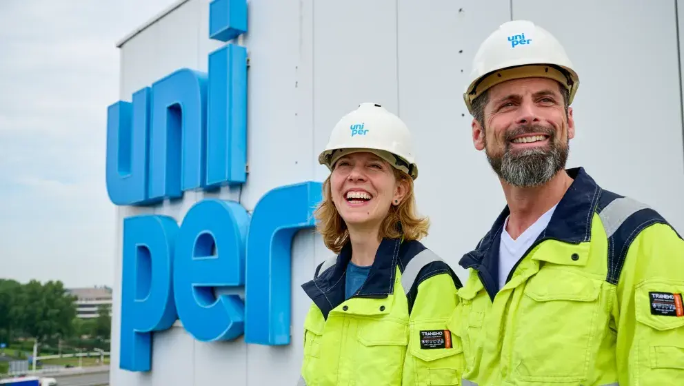 Two smiling Uniper employees, a woman and a man, standing in front of a large Uniper logo. Both are wearing safety helmets and yellow work jackets