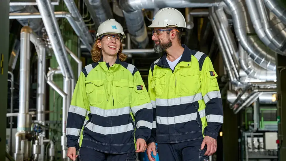 Two Uniper employees, a man and a woman, walking together inside a facility. They are smiling and wearing safety helmets and yellow work jackets with reflective strips.