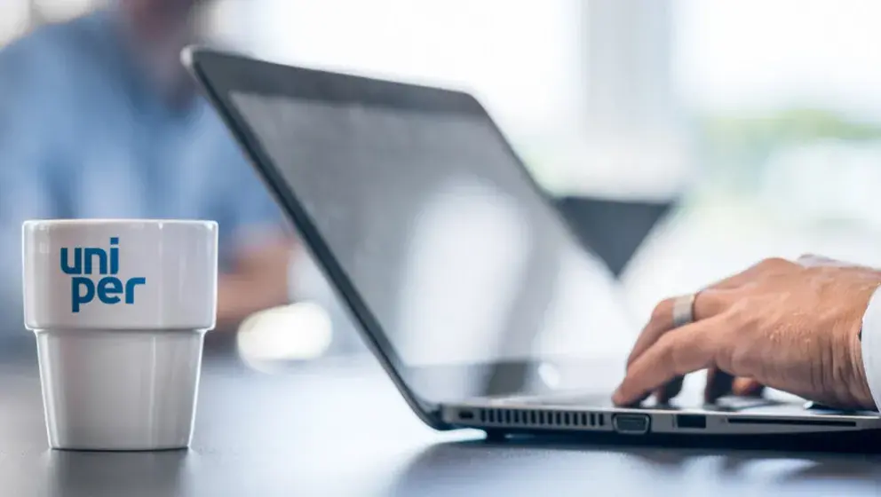A Uniper-branded coffee cup placed next to a laptop on a desk, with a person's hand visible typing on the laptop keyboard