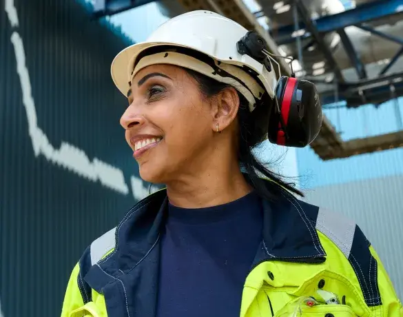 A worker wearing a hard hat and ear protection, smiling while standing outdoors at an industrial site.