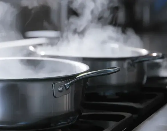 Two stainless steel pots on a stovetop, with steam rising from them, indicating that the contents are being heated or boiled.