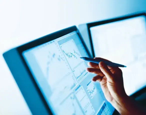 A hand holding a pen and pointing at financial data displayed on computer screens showing graphs and charts