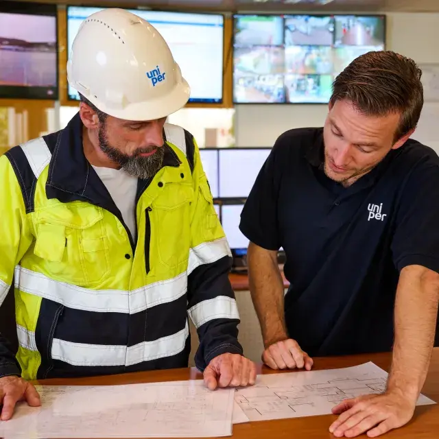 Two Uniper employees in a control room reviewing technical plans, with monitors displaying various operational views in the background.