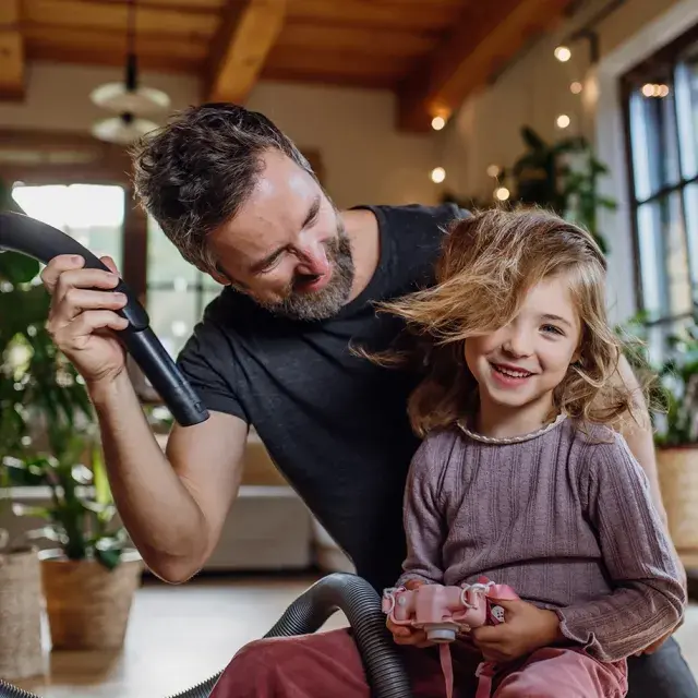 A father playfully using a vacuum hose to blow air into his daughter's hair while his son laughs nearby, creating a fun family moment indoors.
