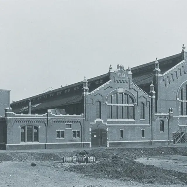 A technician in safety gear and a hard hat holds up a beaker with a pink liquid, carefully inspecting it in a laboratory setting.