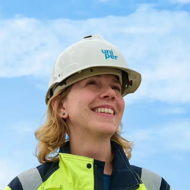 A smiling worker wearing a Uniper-branded hard hat and high-visibility jacket, standing outdoors against a bright blue sky.