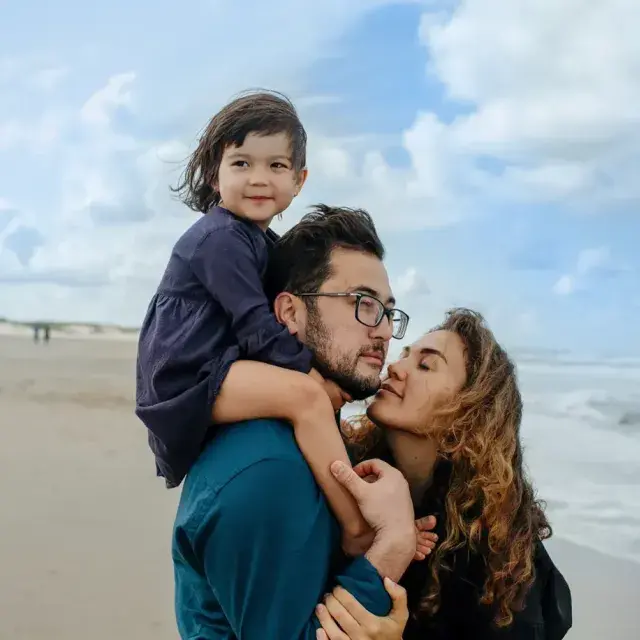 A family standing on a beach, with a young child on the father's shoulders and the mother kissing the father's cheek, as they enjoy a day by the sea.