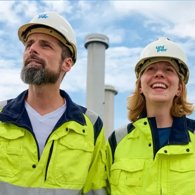 Two Uniper workers in high-visibility clothing and safety helmets standing outdoors, smiling, with industrial chimneys in the background.