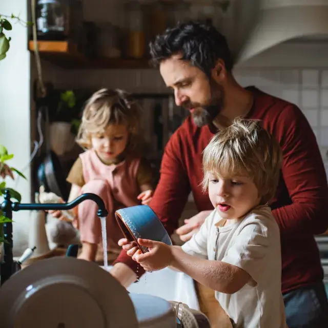 A father and his two young children washing dishes together in a cozy, plant-filled kitchen, symbolizing sustainable living and family care.